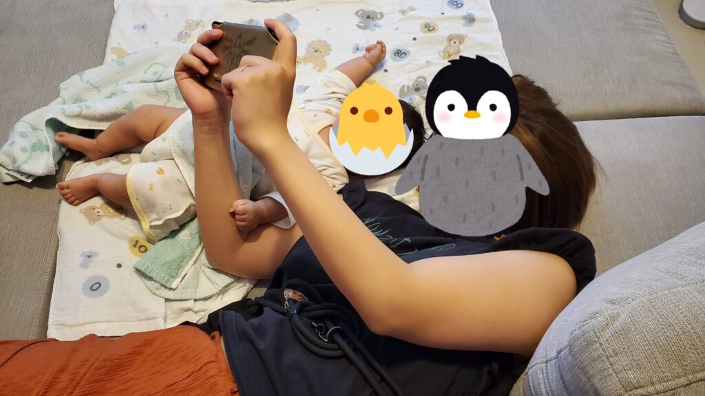 寝っ転がってNintendo Switchで遊ぶ妻とその横で画面を見ている生後2か月の娘